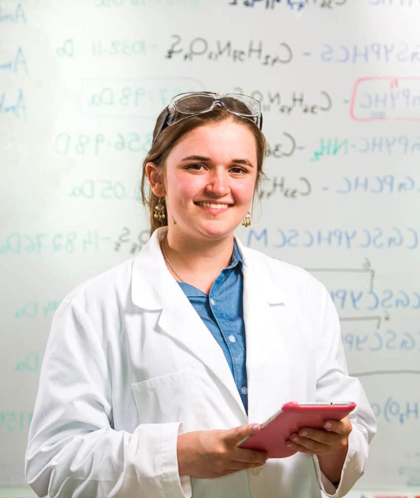 A student in a lab coat in front of a white board filled with chemistry codes. 